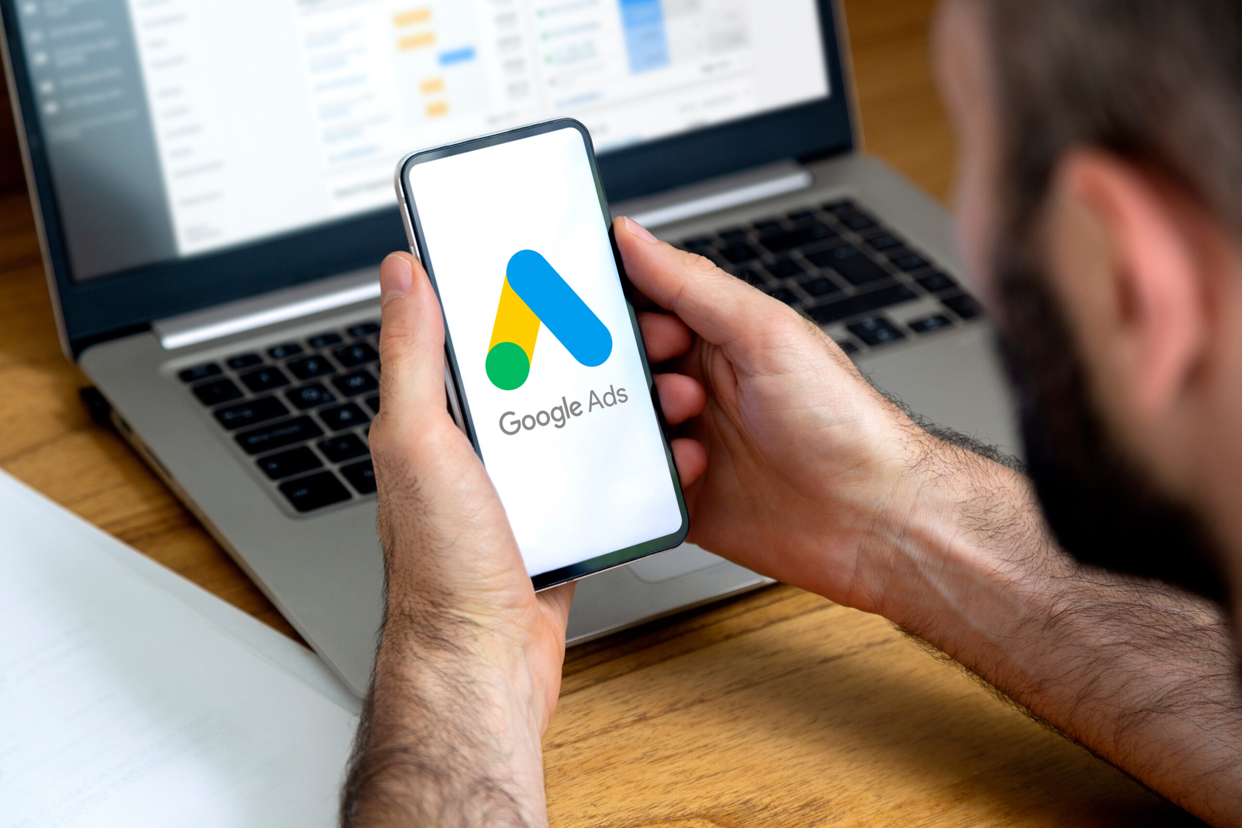 Man using a mobile phone with Google Ads on the screen and in the background a computer showing ad statistics. Rosario, Argentina – May 23, 2024. google ads performance max setup